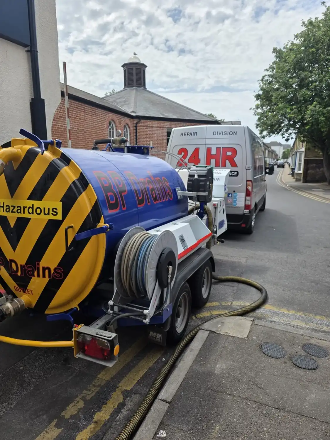 Blue waste removal truck with “BP Drains” signage parked on a street, with a hose extending to the ground. Building and tree in background.